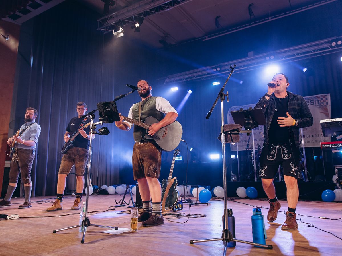 Die ganze Band in Lederhosen auf der Bühne der Zerbster Schloss-Wiesn, Sänger am Mikrofon, blaues Bühnenlicht